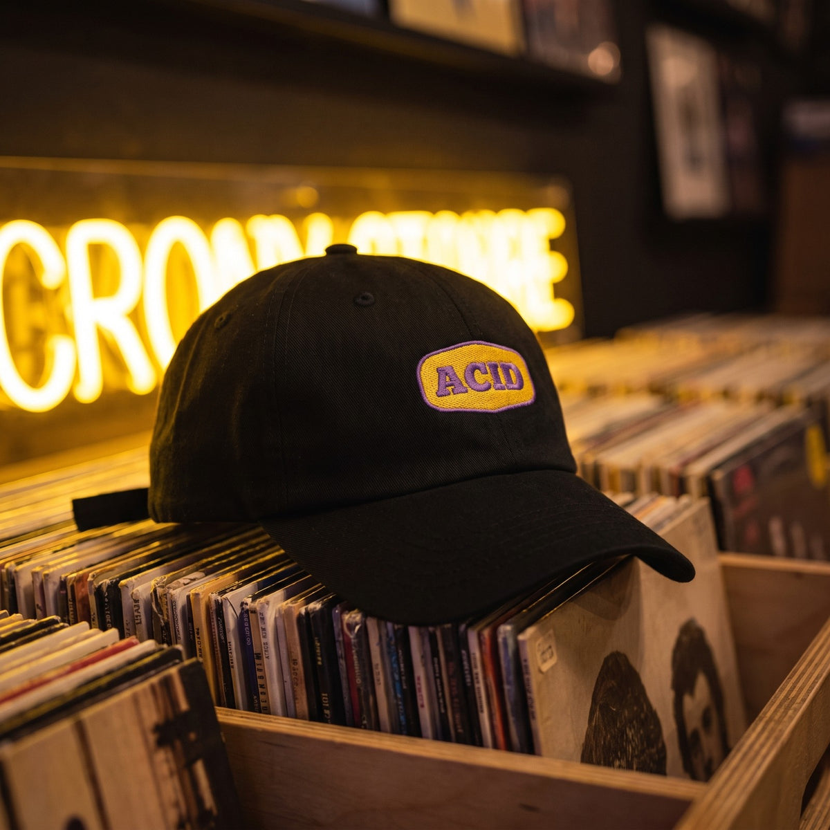 Casquette baseball noire brodée d'un patch jaune aux coins arrondis avec ACID en violet posée sur un bac de disquaire rempli de vinyles avec une enseigne néon jaune derrière - CHT'M Boutique