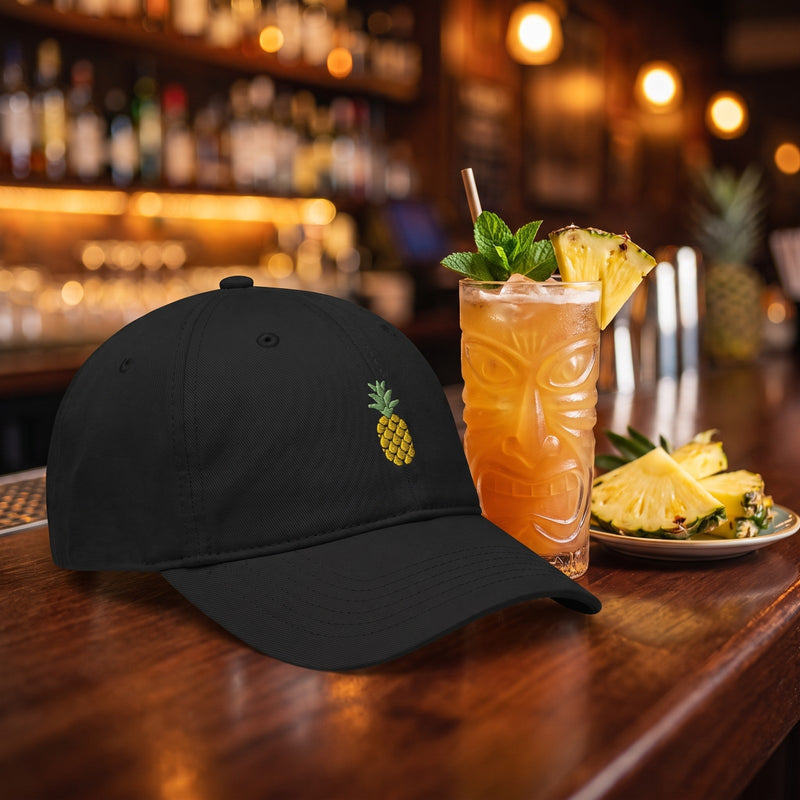 Casquette baseball noire brodée d'un ananas doré posée sur un comptoir de bar à côté d'un cocktail tropical et de tranches d'ananas - CHT'M Boutique