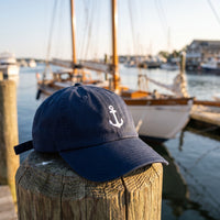 Casquette baseball bleu marine brodée d'une ancre blanche posée sur un poteau de ponton en bois dans un port avec un voilier flou derrière, lumière fraîche du matin - CHT'M Boutique
