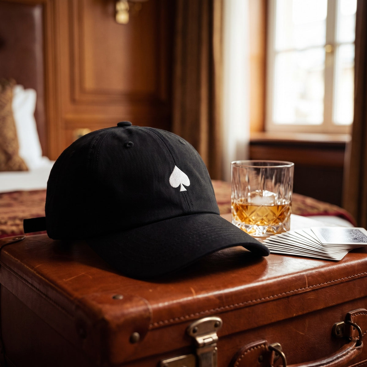 Casquette baseball noire brodée d'un as de pique blanc posée sur une valise en cuir dans une suite d'hôtel à côté d'un verre de whisky et d'un jeu de cartes - CHT'M Boutique