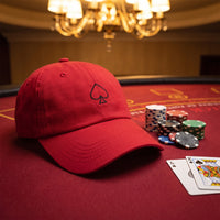 Casquette baseball rouge brodée d'un pique noir au trait posée sur un tapis rouge de blackjack à côté d'une pile de jetons et de deux cartes, lumière chaude - CHT'M Boutique