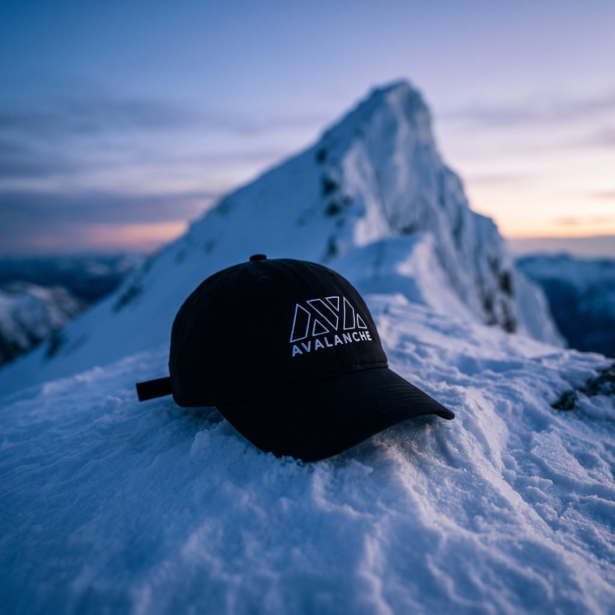 Casquette baseball noire brodée du logo AVALANCHE en blanc au trait posée sur une crête enneigée à l'aube devant un pic glacé escarpé, lumière bleutée froide - CHT'M Boutique