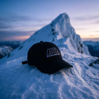 Casquette baseball noire brodée du logo AVALANCHE en blanc au trait posée sur une crête enneigée à l'aube devant un pic glacé escarpé, lumière bleutée froide - CHT'M Boutique