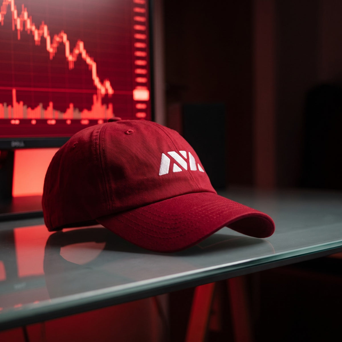 Casquette baseball rouge canneberge brodée du logo AVAX triangle en blanc posée sur un bureau en verre à côté d'un écran de graphique en bougies rougeoyant, pièce sombre - CHT'M Boutique