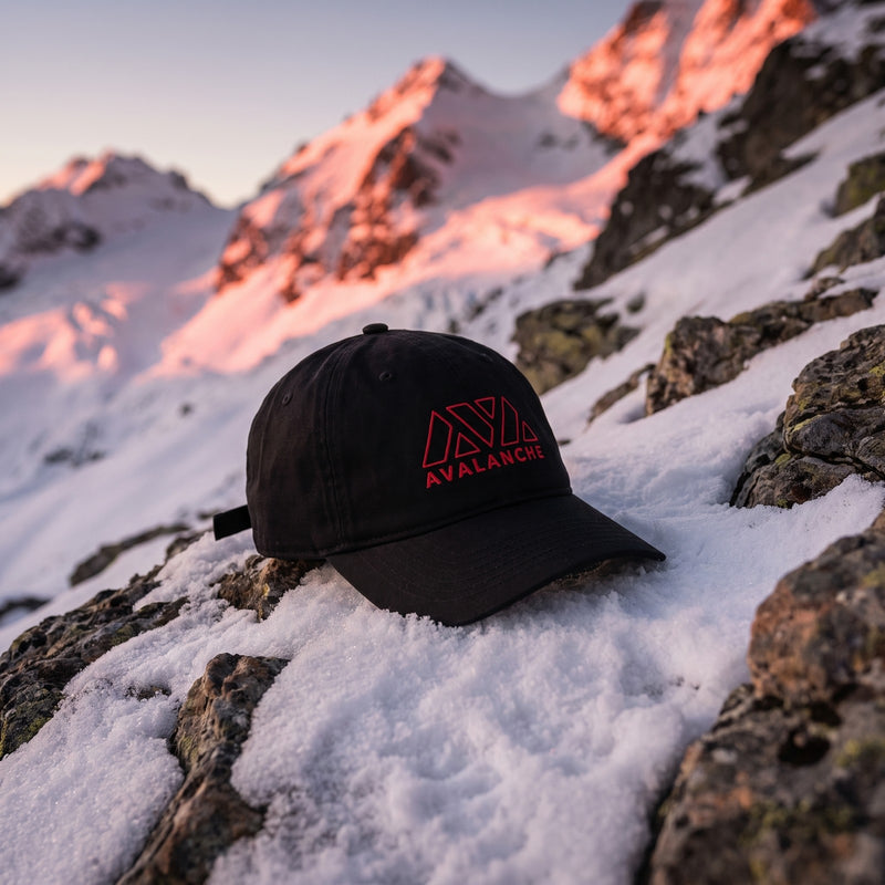 Casquette baseball noire brodée du logo AVALANCHE en rouge au trait posée sur une pente rocheuse après une petite coulée de neige, avec une lueur rouge sur les sommets - CHT'M Boutique