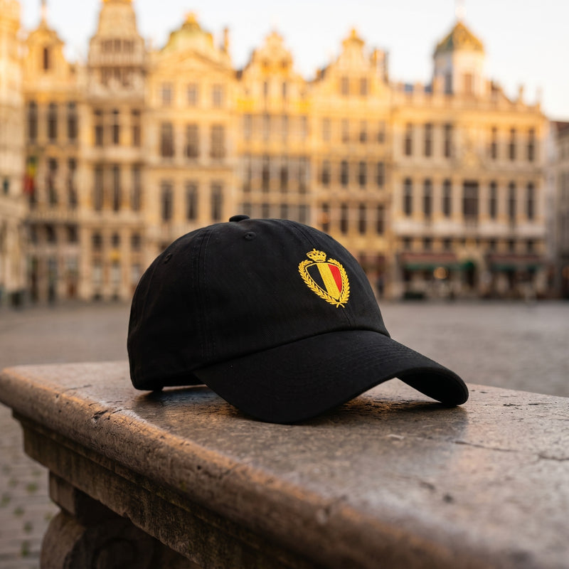 Casquette baseball noire brodée d'un blason belge couronné doré aux couleurs noir, jaune et rouge posée sur un banc en pierre devant la Grand-Place de Bruxelles à l'heure dorée, arrière-plan flou - CHT'M Boutique