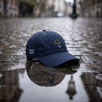 Casquette baseball bleu marine brodée d'un petit parapluie jaune au trait avec un texte blanc sur le côté posée sur un pavé mouillé avec des gouttes de pluie et une lumière grise douce, une flaque reflétant un lampadaire belge - CHT'M Boutique