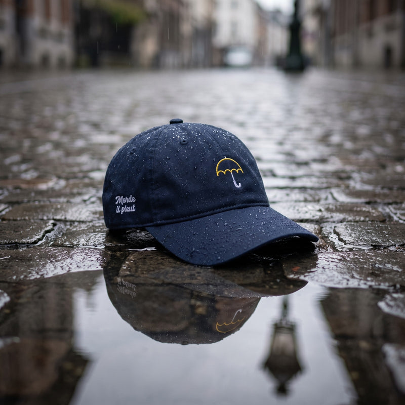 Casquette baseball bleu marine brodée d'un petit parapluie jaune au trait avec un texte blanc sur le côté posée sur un pavé mouillé avec des gouttes de pluie et une lumière grise douce, une flaque reflétant un lampadaire belge - CHT'M Boutique
