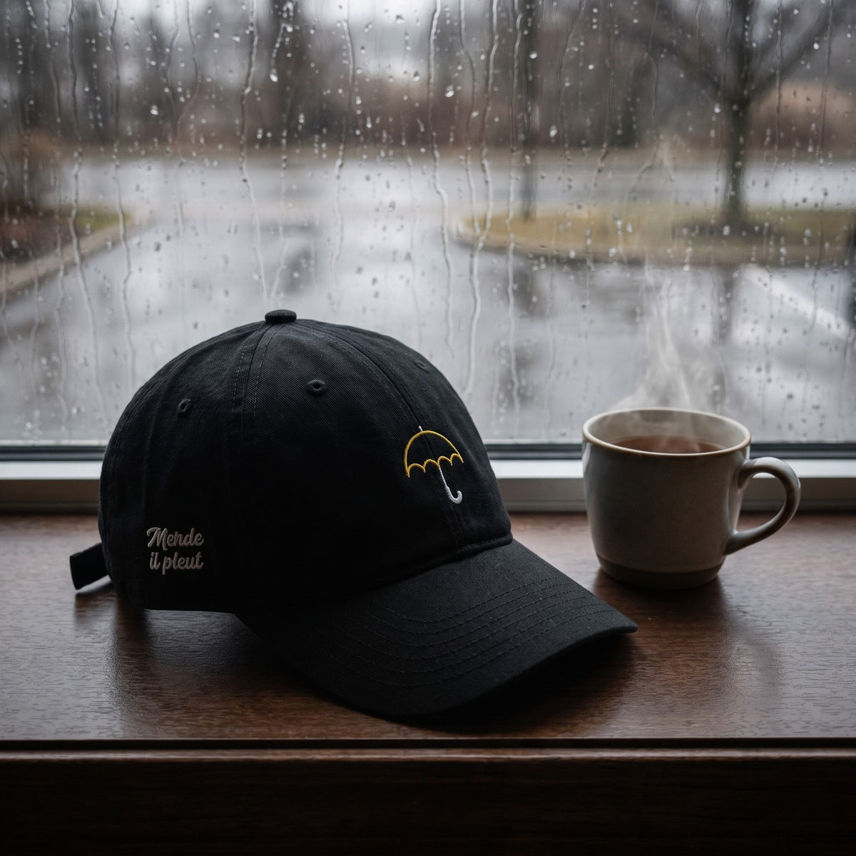 Casquette baseball noire brodée d'un petit parapluie jaune au trait avec un texte blanc sur le côté posée sur un rebord de fenêtre avec de la pluie qui ruisselle sur la vitre derrière et une tasse de thé, lumière grise cosy - CHT'M Boutique