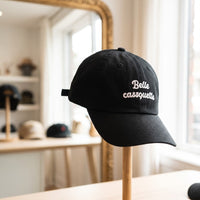 Casquette baseball noire brodée Belle casquette en écriture blanche sur deux lignes posée sur un présentoir à chapeaux dans une boutique lumineuse avec un miroir derrière, lumière douce du jour - CHT'M Boutique