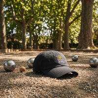 Casquette baseball noire brodée d'un patch gris avec une couronne dorée et BOULE KING en blanc posée sur un terrain de pétanque en gravier à l'ombre de platanes à côté de boules en acier et d'un cochonnet, lumière tamisée - CHT'M Boutique