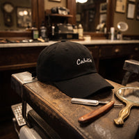 Casquette baseball noire brodée Calvitie en écriture manuscrite blanche posée sur l'accoudoir d'un fauteuil de barbier vintage à côté d'un rasoir coupe-chou et d'un miroir à main - CHT'M Boutique