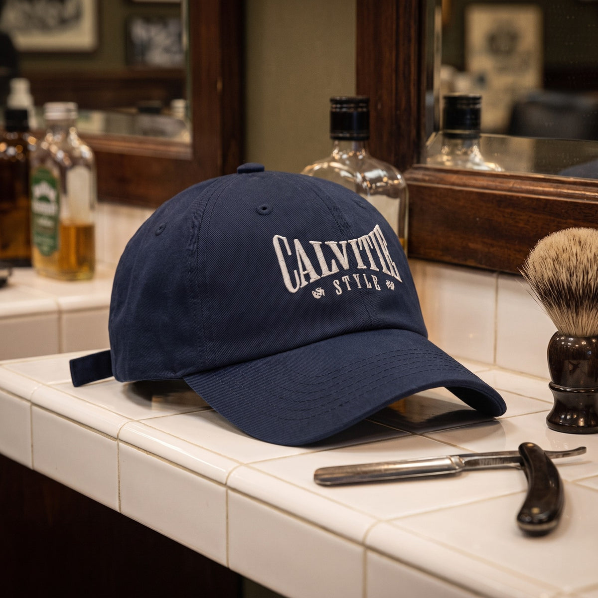 Casquette baseball bleu marine brodée CALVITIE STYLE en arc, lettres gris clair posée sur une étagère carrelée de blanc dans un salon de barbier à côté d'un rasoir coupe-chou et d'un blaireau - CHT'M Boutique
