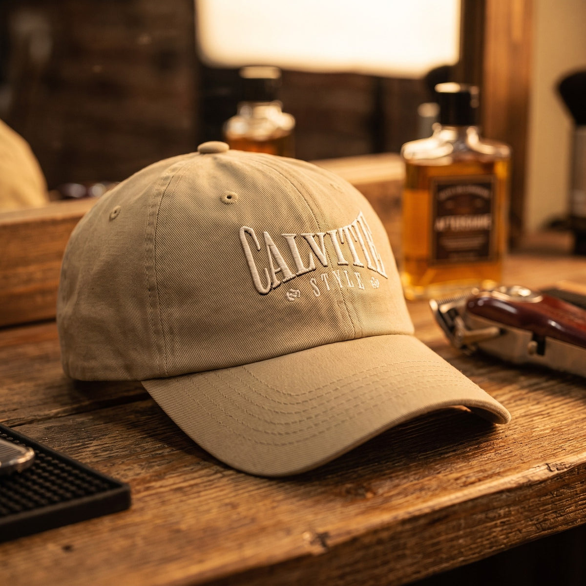 Casquette baseball kaki brodée CALVITIE STYLE en arc, lettres crème posée sur le comptoir d'un salon de barbier à côté d'une tondeuse et d'un flacon de lotion après-rasage sous une lumière chaude - CHT'M Boutique