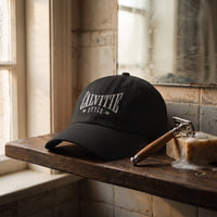 Casquette baseball noire brodée CALVITIE STYLE en arc, lettres gris clair posée sur une étagère de salle de bain en bois sombre à côté d'un rasoir et d'un savon sous une lumière douce - CHT'M Boutique