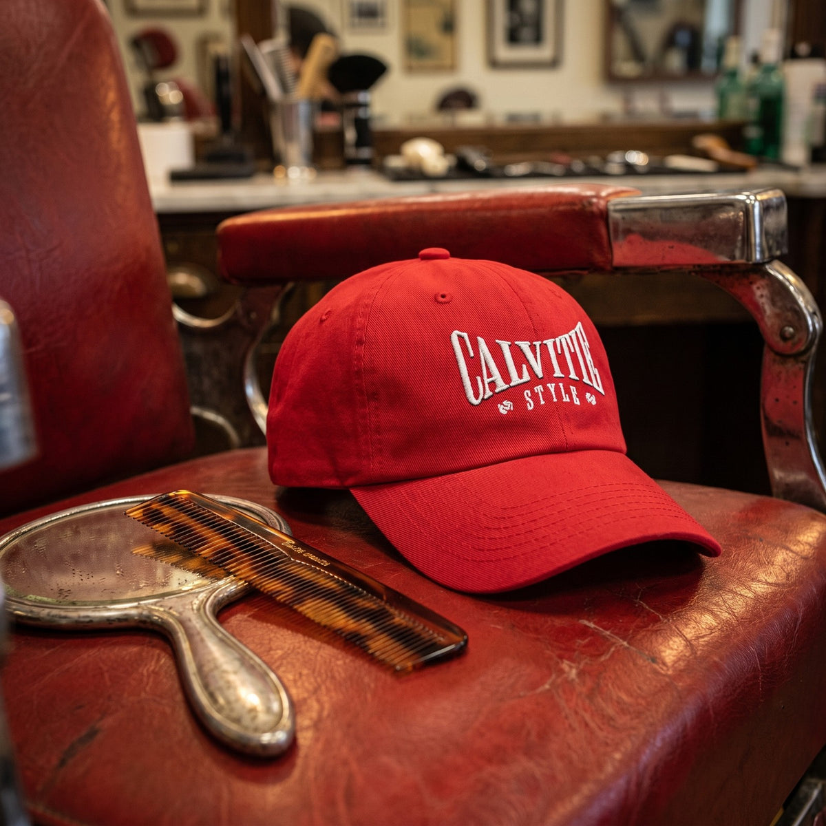 Casquette baseball rouge brodée CALVITIE STYLE en arc, lettres blanches posée sur l'accoudoir d'un fauteuil de barbier vintage en cuir rouge à côté d'un peigne et d'un miroir - CHT'M Boutique