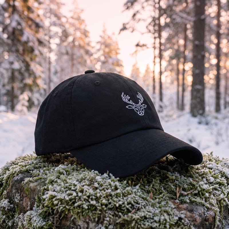 Casquette baseball noire brodée d'une tête de cerf origami géométrique blanche posée sur une pierre moussue dans une forêt enneigée à l'aube, lumière rose pâle - CHT'M Boutique