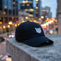 Casquette baseball noire brodée d'une petite tête de chat géométrique blanche posée sur le rebord d'un toit en ville à l'heure bleue avec des lumières floues derrière - CHT'M Boutique