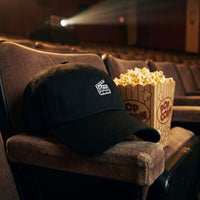 Casquette baseball noire brodée d'un petit clap de cinéma blanc posée sur l'accoudoir d'un siège de salle de cinéma à côté d'un seau de popcorn sous la lumière tamisée du projecteur - CHT'M Boutique