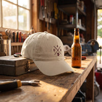 Casquette baseball oyster brodée du monogramme DAD bordeaux avec une chope de bière posée sur un établi de garage à côté d'une boîte à outils et d'une bière fraîche sous une lumière chaude d'après-midi - CHT'M Boutique