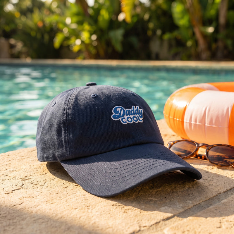Casquette baseball bleu marine brodée Daddy COOL en écriture rétro bleue cernée de blanc posée sur le bord d'une piscine en été à côté de lunettes de soleil et d'une bouée, lumière vive - CHT'M Boutique
