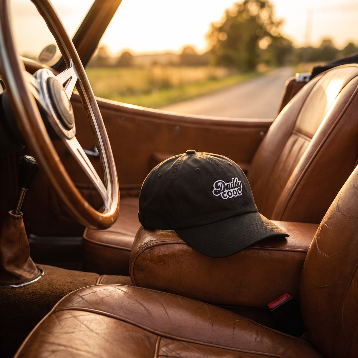 Casquette baseball noire brodée Daddy COOL en écriture rétro blanche cernée de gris posée sur le siège en cuir d'une décapotable vintage avec le volant en vue, lumière dorée de coucher de soleil - CHT'M Boutique