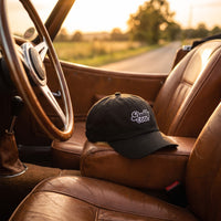 Casquette baseball noire brodée Daddy COOL en écriture rétro blanche cernée de gris posée sur le siège en cuir d'une décapotable vintage avec le volant en vue, lumière dorée de coucher de soleil - CHT'M Boutique