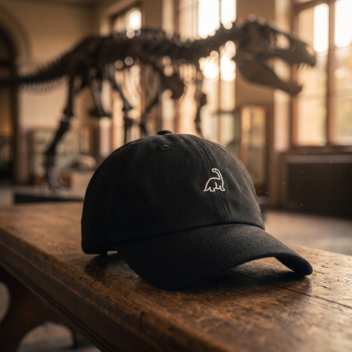 Casquette baseball noire brodée d'un petit dinosaure brontosaure blanc au trait posée sur un banc de musée devant un squelette de dinosaure flou - CHT'M Boutique