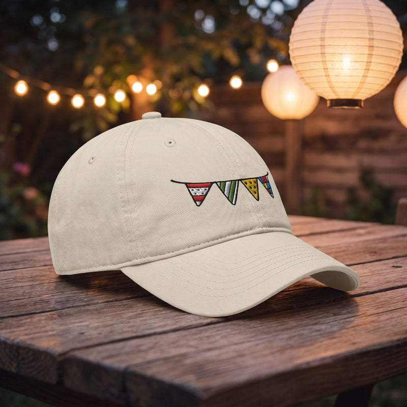 Casquette baseball oyster brodée d'une guirlande de petits fanions colorés posée sur une table de fête en plein air avec des guirlandes lumineuses et des lampions en papier au crépuscule - CHT'M Boutique