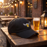 Casquette baseball noire brodée d'une chope de bière dorée avec des ailes blanches posée sur une table en bois d'une terrasse de brasserie belge à côté d'une bière mousseuse sous la lumière chaude d'une lanterne - CHT'M Boutique
