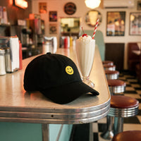 Casquette baseball noire brodée d'un petit smiley jaune posée sur le comptoir d'un diner américain rétro à côté d'un milkshake - CHT'M Boutique