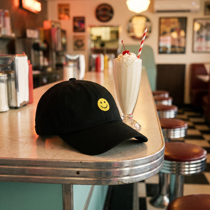 Casquette baseball noire brodée d'un petit smiley jaune posée sur le comptoir d'un diner américain rétro à côté d'un milkshake - CHT'M Boutique