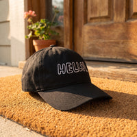Casquette baseball noire brodée HELLO! en lettres blanches dessinées au trait posée sur un paillasson devant une porte d'entrée avec une petite plante en pot, lumière du matin - CHT'M Boutique