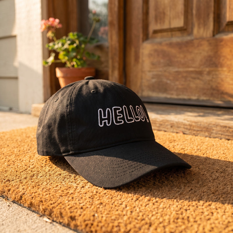 Casquette baseball noire brodée HELLO! en lettres blanches dessinées au trait posée sur un paillasson devant une porte d'entrée avec une petite plante en pot, lumière du matin - CHT'M Boutique