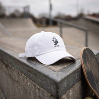 Casquette baseball blanche brodée d'un petit fantôme noir sur un skateboard avec le texte HEY BOO posée sur un muret en béton de skatepark à côté d'une planche de skate sous une lumière douce de ciel couvert - CHT'M Boutique