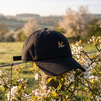 Casquette baseball noire brodée d'une petite hirondelle dorée et rouge posée sur un piquet de clôture dans une campagne printanière avec des fleurs, lumière douce du matin - CHT'M Boutique