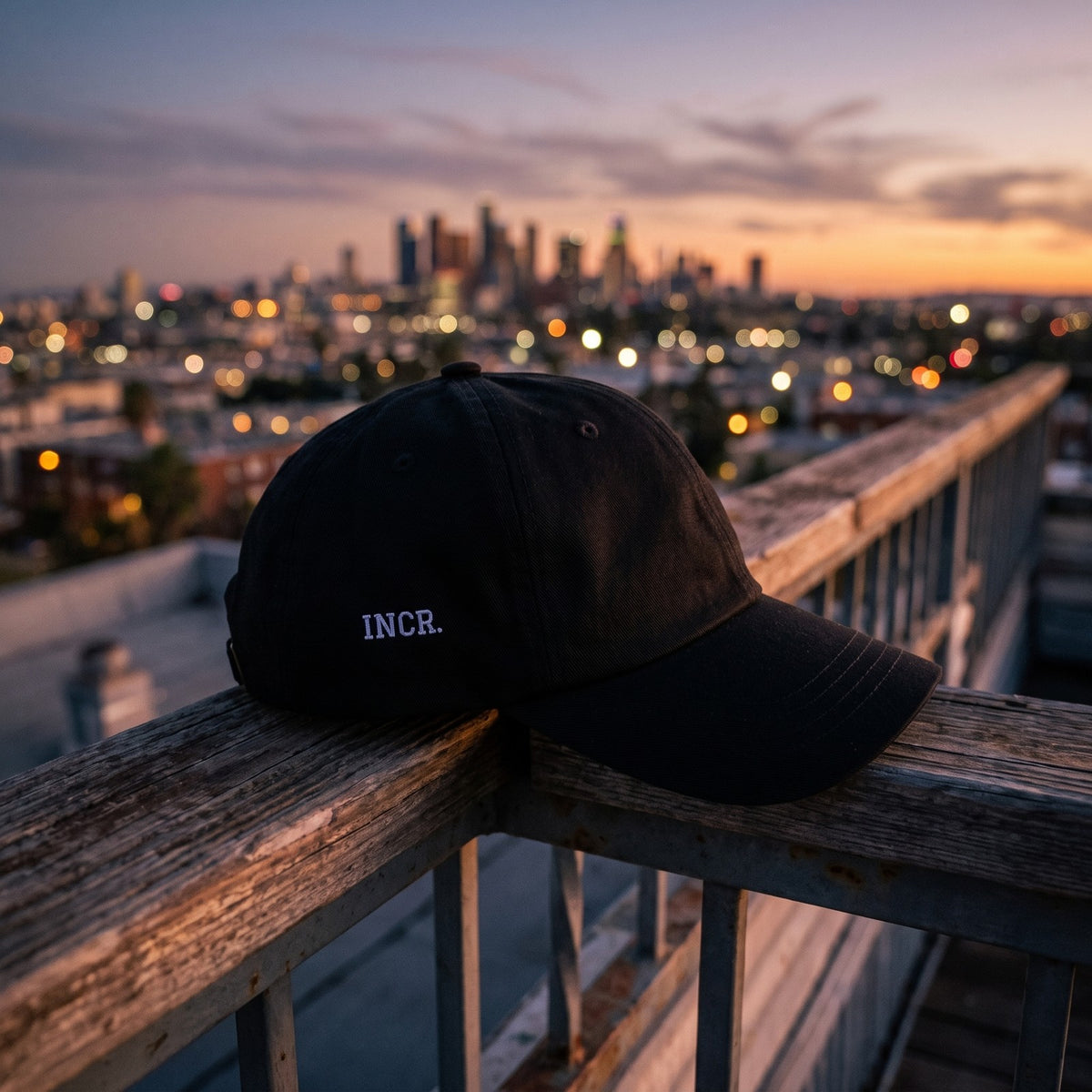 Casquette baseball noire brodée INCR. en petit blanc sur le côté posée sur la rambarde d'un toit-terrasse au crépuscule devant les lumières d'une ville - CHT'M Boutique