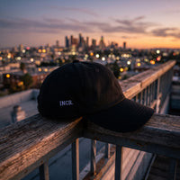 Casquette baseball noire brodée INCR. en petit blanc sur le côté posée sur la rambarde d'un toit-terrasse au crépuscule devant les lumières d'une ville - CHT'M Boutique