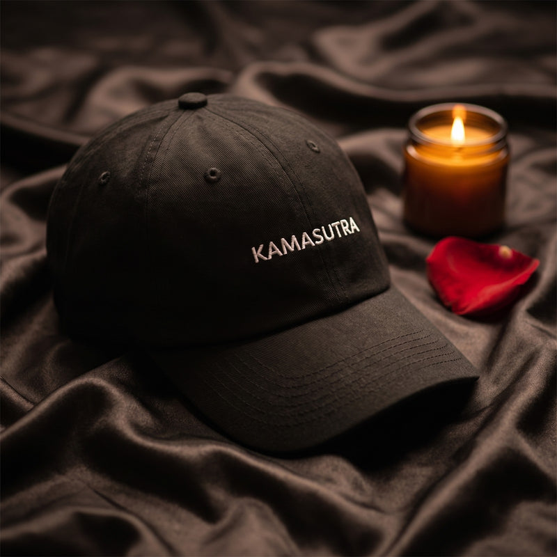 Casquette baseball noire brodée KAMASUTRA en petites capitales blanches posée sur des draps de satin sombres froissés avec un pétale de rose et une bougie, lumière chaude tamisée - CHT'M Boutique