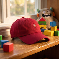 Casquette baseball rouge canneberge brodée d'un petit monstre rouge de dessin animé aux bras levés posée sur l'étagère d'une chambre d'enfant parmi des cubes colorés et une peluche, lumière chaude et joyeuse - CHT'M Boutique