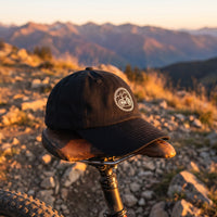 Casquette baseball noire brodée d'un badge rond blanc avec des montagnes et un VTT posée sur la selle d'un VTT à un point de vue rocheux d'un sentier au lever du soleil - CHT'M Boutique