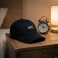 Casquette baseball noire brodée OFF. en blanc posée sur une table de chevet à côté d'un réveil éteint et d'une lampe, lumière calme de nuit - CHT'M Boutique