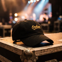 Casquette baseball noire brodée Orelsan en écriture dorée posée sur un flight case en coulisses avec des projecteurs de scène flous derrière - CHT'M Boutique