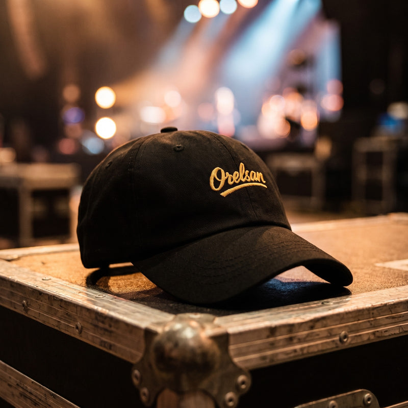 Casquette baseball noire brodée Orelsan en écriture dorée posée sur un flight case en coulisses avec des projecteurs de scène flous derrière - CHT'M Boutique