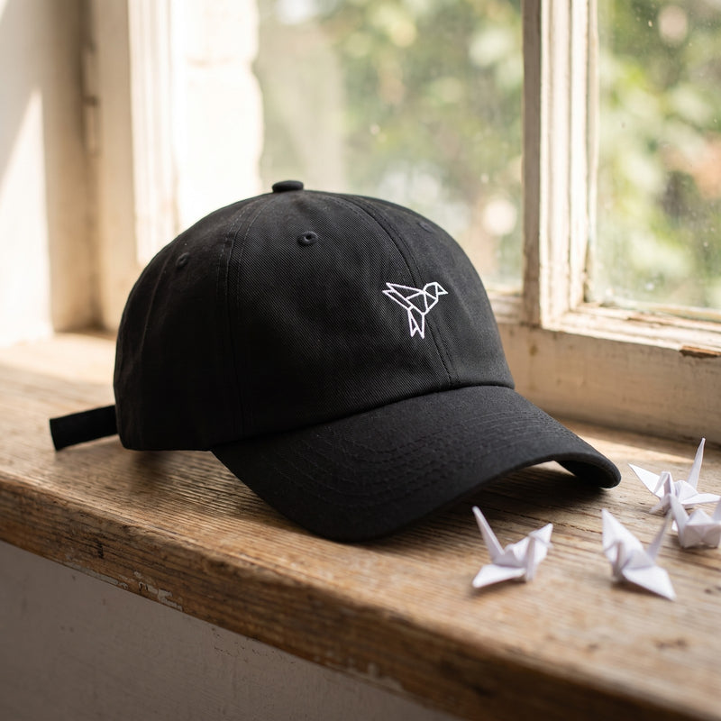 Casquette baseball noire brodée d'un petit oiseau origami géométrique blanc posée sur le rebord d'une fenêtre à côté de grues en papier pliées sous une lumière douce du jour - CHT'M Boutique
