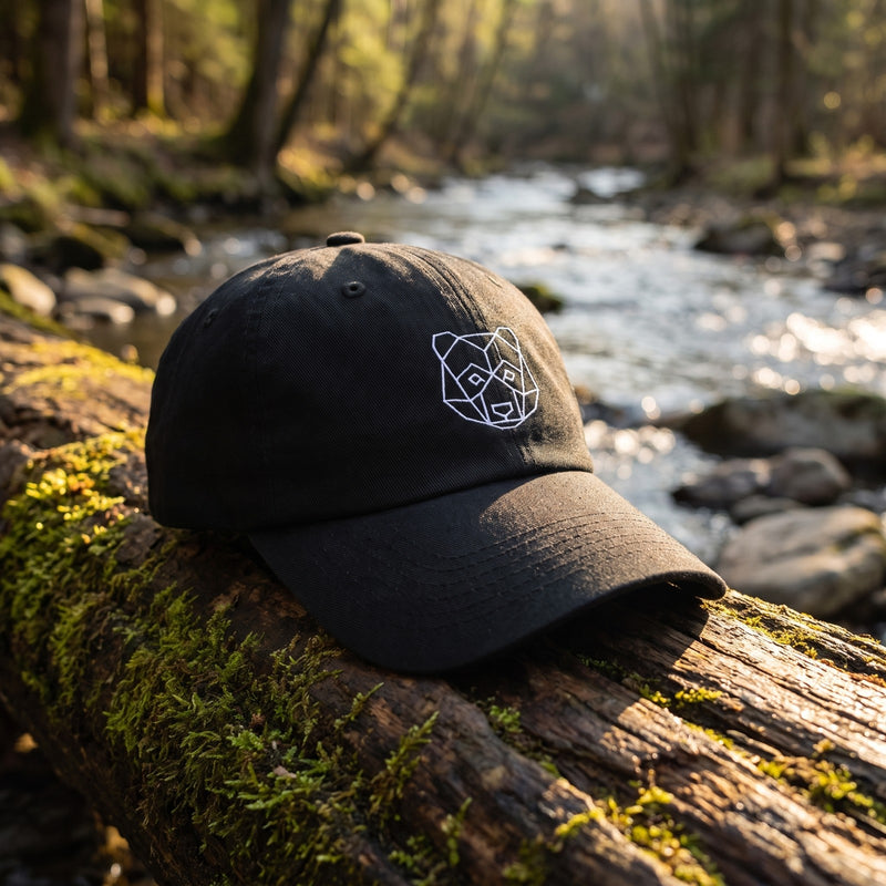 Casquette baseball noire brodée d'une tête d'ours origami géométrique blanche posée sur un tronc couché au bord d'un ruisseau de montagne calme dans la lumière du petit matin - CHT'M Boutique
