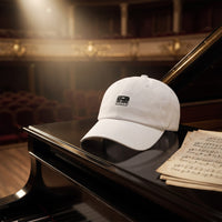 Casquette baseball blanche brodée d'une petite icône de clavier de piano noire posée sur le couvercle fermé d'un piano à queue noir à côté de partitions sous une lumière douce - CHT'M Boutique