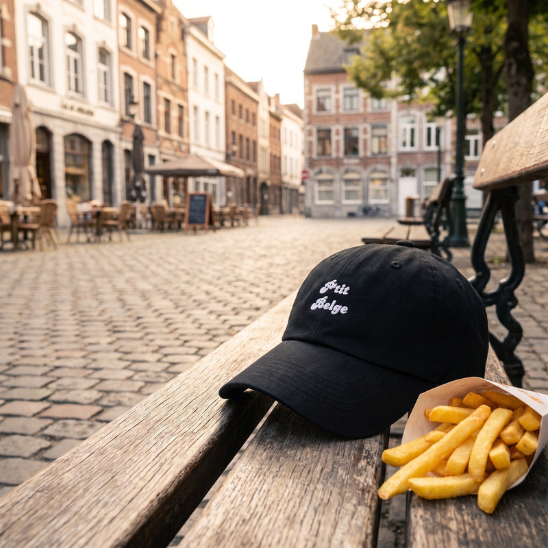 Casquette baseball noire brodée P'tit Belge en écriture rétro blanche sur deux lignes posée sur un banc d'une place de village belge à côté d'un cornet de frites sous une lumière douce d'après-midi - CHT'M Boutique