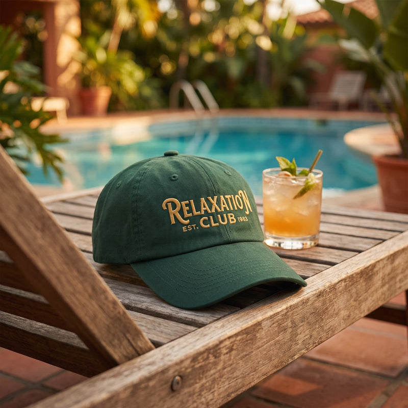 Casquette baseball vert forêt brodée RELAXATION CLUB EST. 1983 en doré posée sur un transat en bois au bord d'une piscine d'un hôtel rétro à côté d'un cocktail sous la lumière chaude de l'après-midi - CHT'M Boutique
