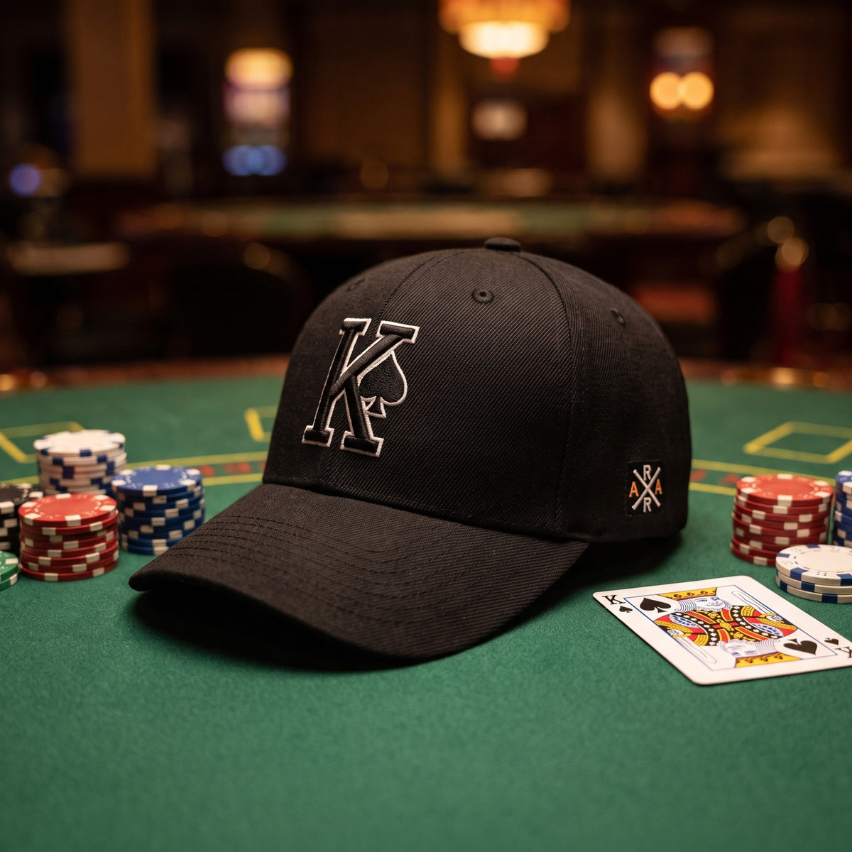 Casquette baseball structurée noire brodée d'un grand K cerné de blanc avec un pique noir posée sur un tapis vert de table de poker à côté de jetons empilés et d'une carte de roi, lumière tamisée de casino - CHT'M Boutique
